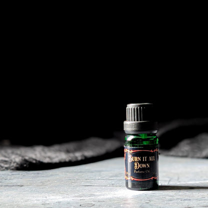 Bottle of 'Burn It All Down' perfume oil on a wooden surface with a dark background