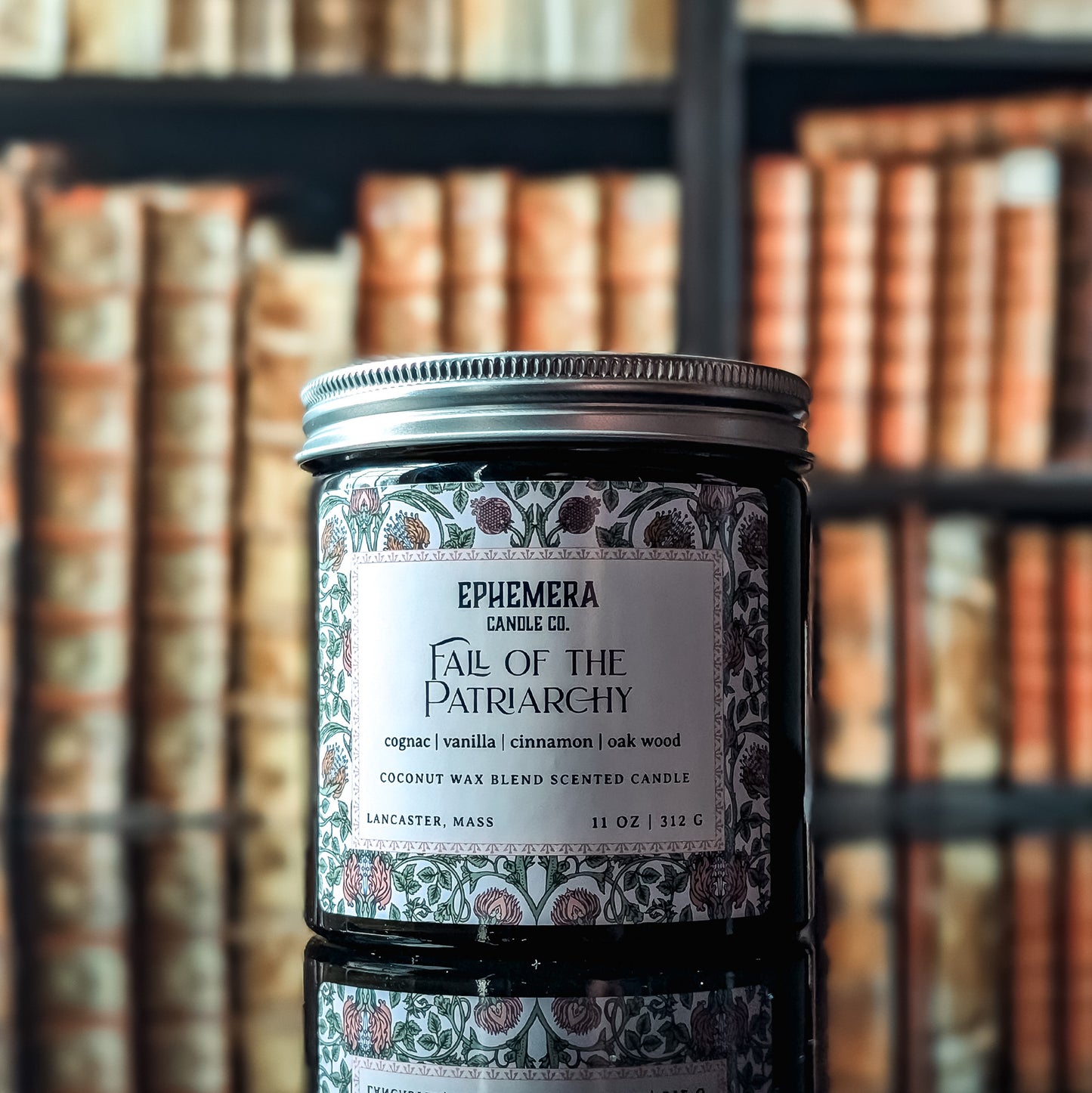 Fall of the Patriarchy candle - green glass jar against a bookshelf background. Cognac, vanilla, cinnamon and oak. 