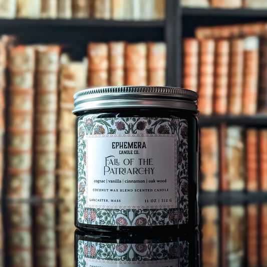 Fall of the Patriarchy candle - green glass jar against a bookshelf background. Cognac, vanilla, cinnamon and oak. 